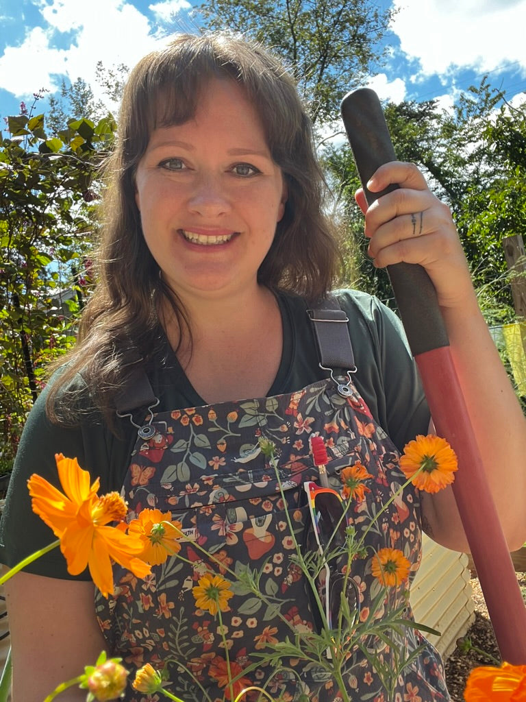 A smiling woman outdoors wearing floral gardening overalls over a dark green shirt, holding a red and black garden rake. Bright orange flowers are in the foreground, with trees and blue sky in the background.