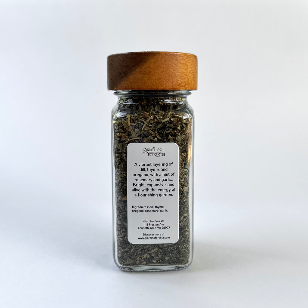 A clear glass spice jar filled with dried herbs, featuring a light brown wooden lid and a white label with black text, set against a white background.