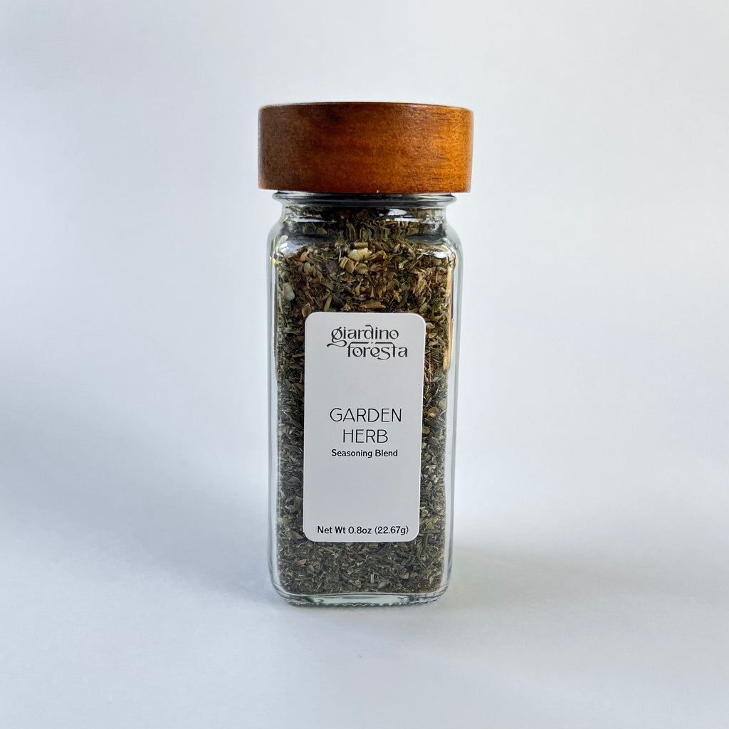 A clear glass spice jar filled with dried herbs, featuring a white label and a medium brown wooden lid, set against a white background.