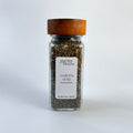 A clear glass spice jar filled with dried herbs, featuring a white label and a medium brown wooden lid, set against a white background.