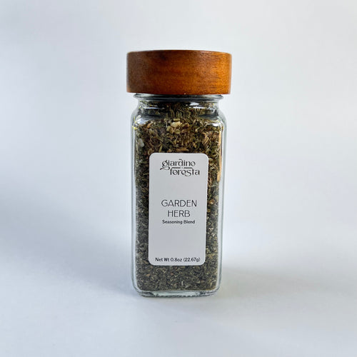 A clear glass spice jar filled with dried herbs, featuring a white label and a medium brown wooden lid, set against a white background.