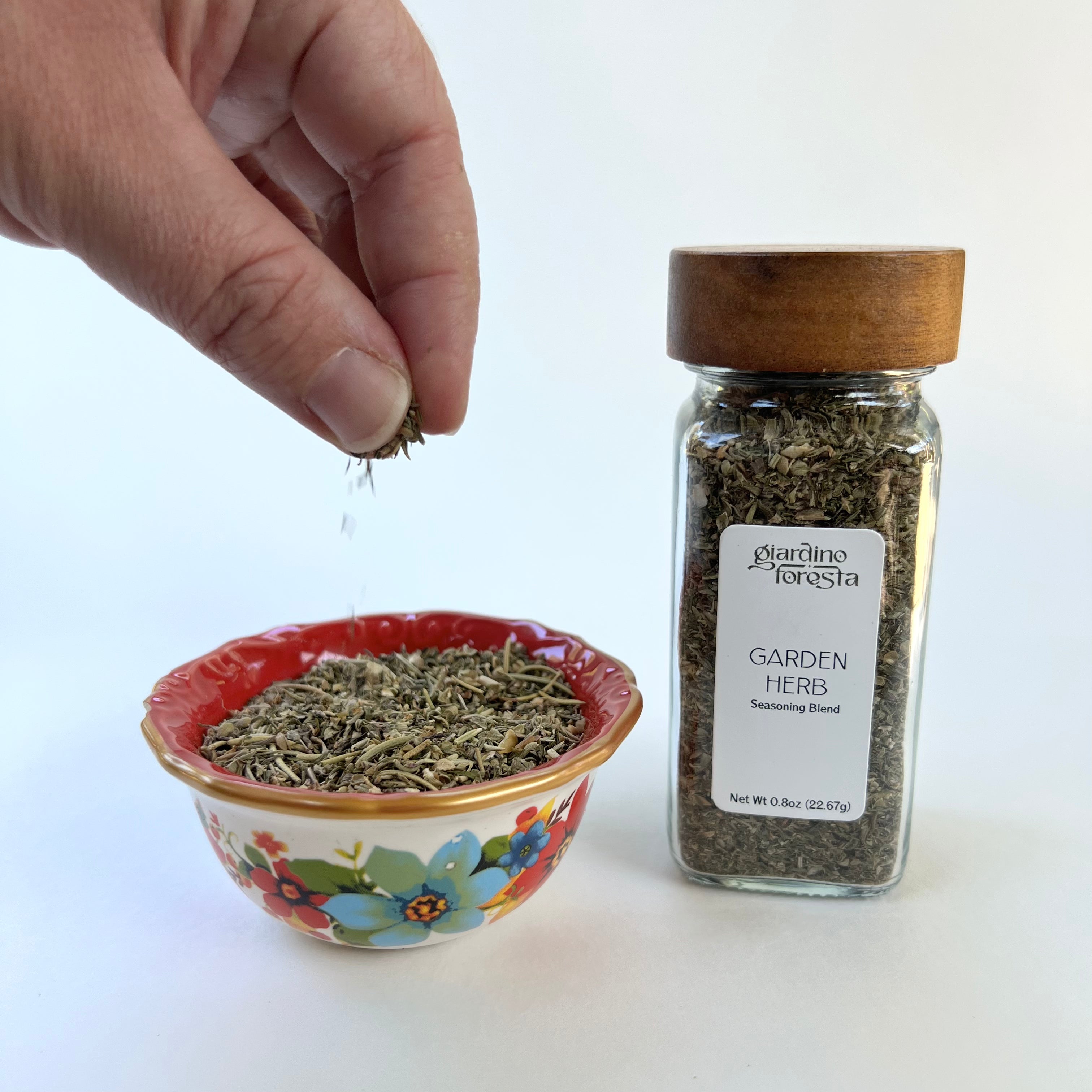 A hand sprinkles dried herbs over a small floral bowl next to a clear glass spice jar with a wooden lid and a white label reading 'Giardino Foresta Garden Herb Seasoning Blend'.