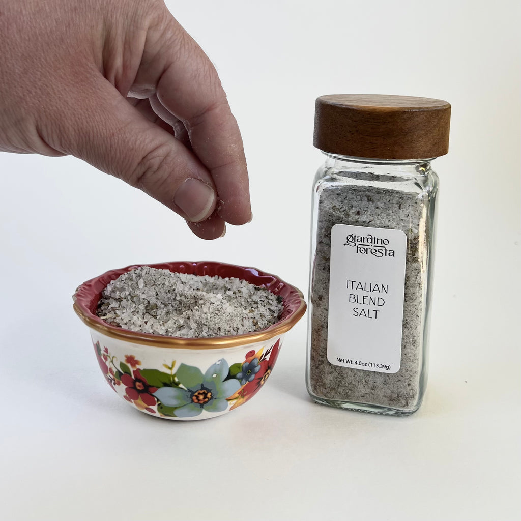 A hand is shown sprinkling salt next to a small floral bowl filled with coarse salt and a glass bottle of Giardino Foresta Italian Blend Salt on a white background.