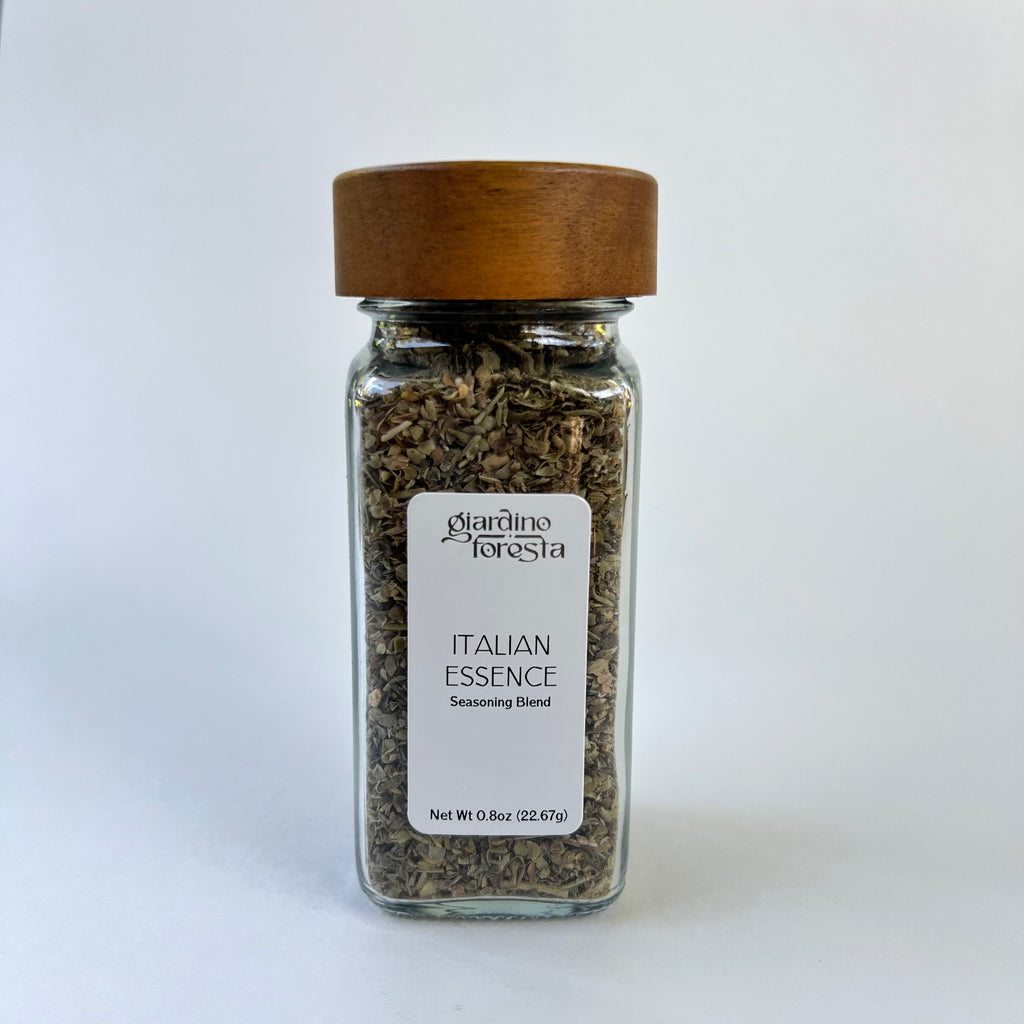 A clear glass spice jar filled with dried herbs, featuring a white label that reads 'Giardino Foresta Italian Essence Seasoning Blend' and a medium brown wooden lid, set against a white background.