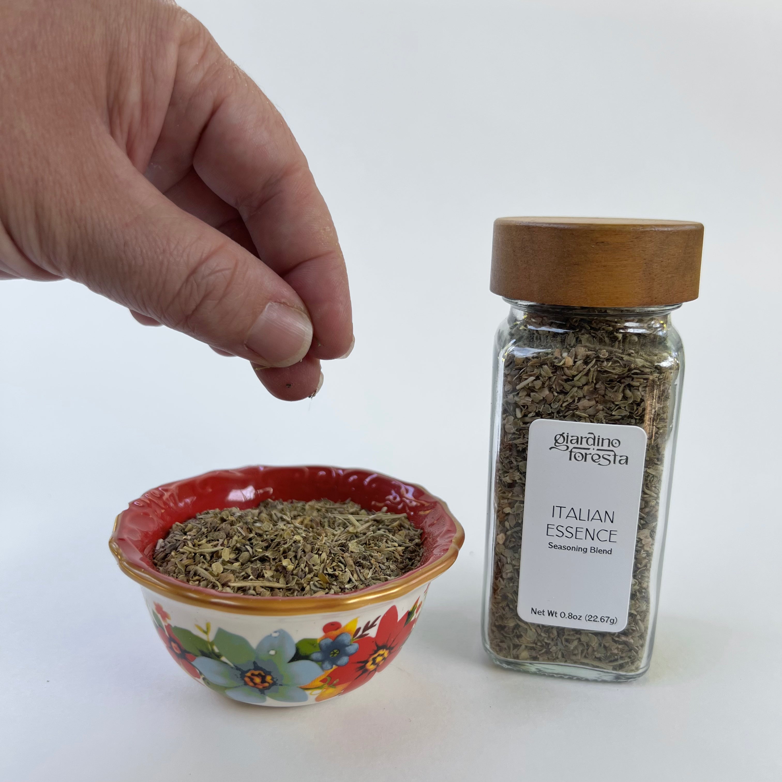A hand is shown sprinkling dried herbs into a small floral bowl next to a glass jar of Giardino Foresta Italian Essence Seasoning Blend on a white background.