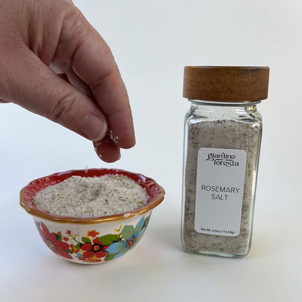 A hand sprinkles seasoning salt into a small floral ceramic bowl next to a glass spice jar labeled 'Giardino Foresta Rosemary Salt' with a wooden lid, on a white background.
