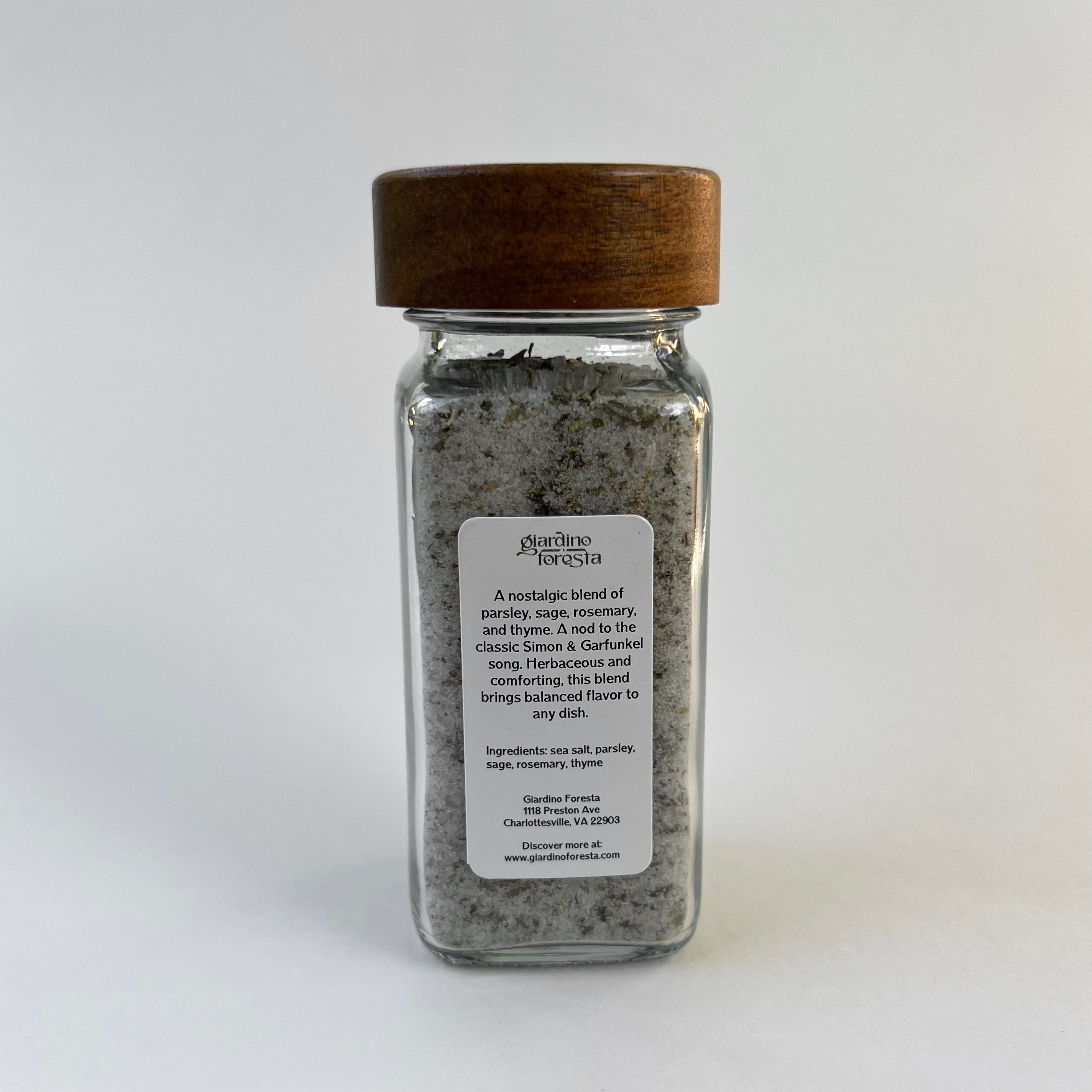 A clear glass spice jar with a dark brown wooden lid, containing seasoning salt. The jar has a white label with black text. The background is solid white.