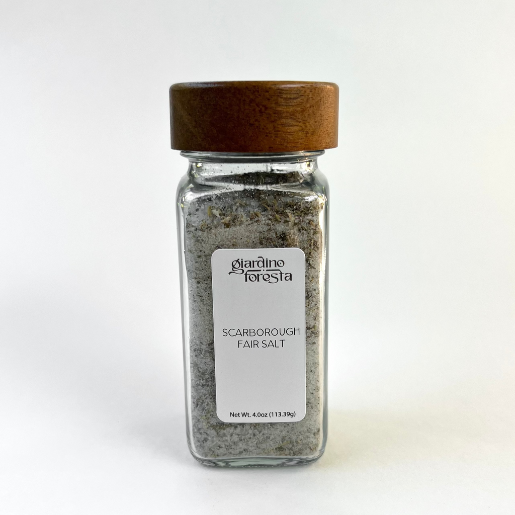 A clear glass spice jar filled with seasoning salt, featuring a medium brown wooden lid and a white label that reads 'Giardino Foresta Scarborough Fair Salt'. The jar is centered against a clean white background.