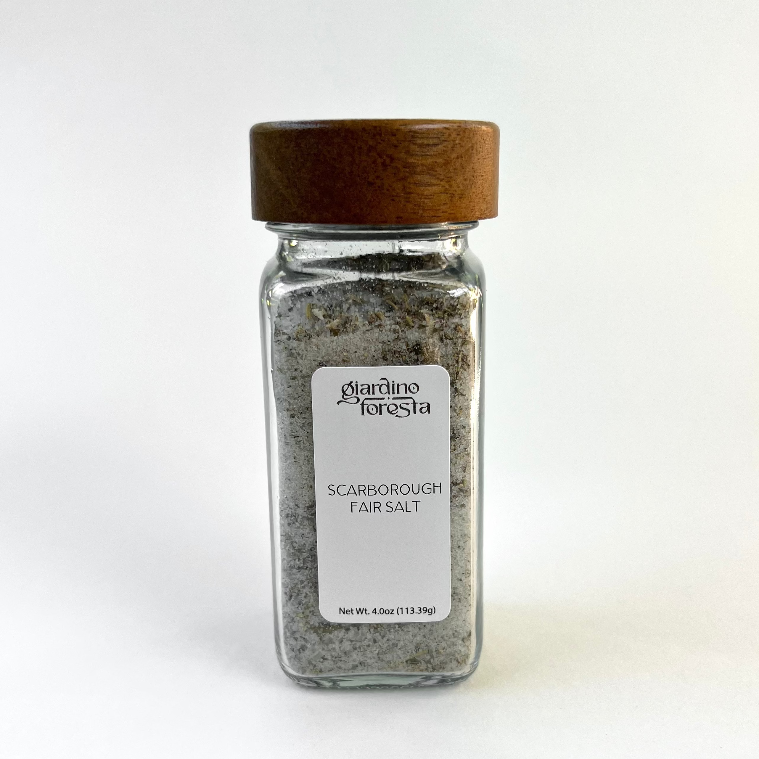 A clear glass spice jar filled with seasoning salt, featuring a medium brown wooden lid and a white label that reads 'Giardino Foresta Scarborough Fair Salt'. The jar is centered against a clean white background.