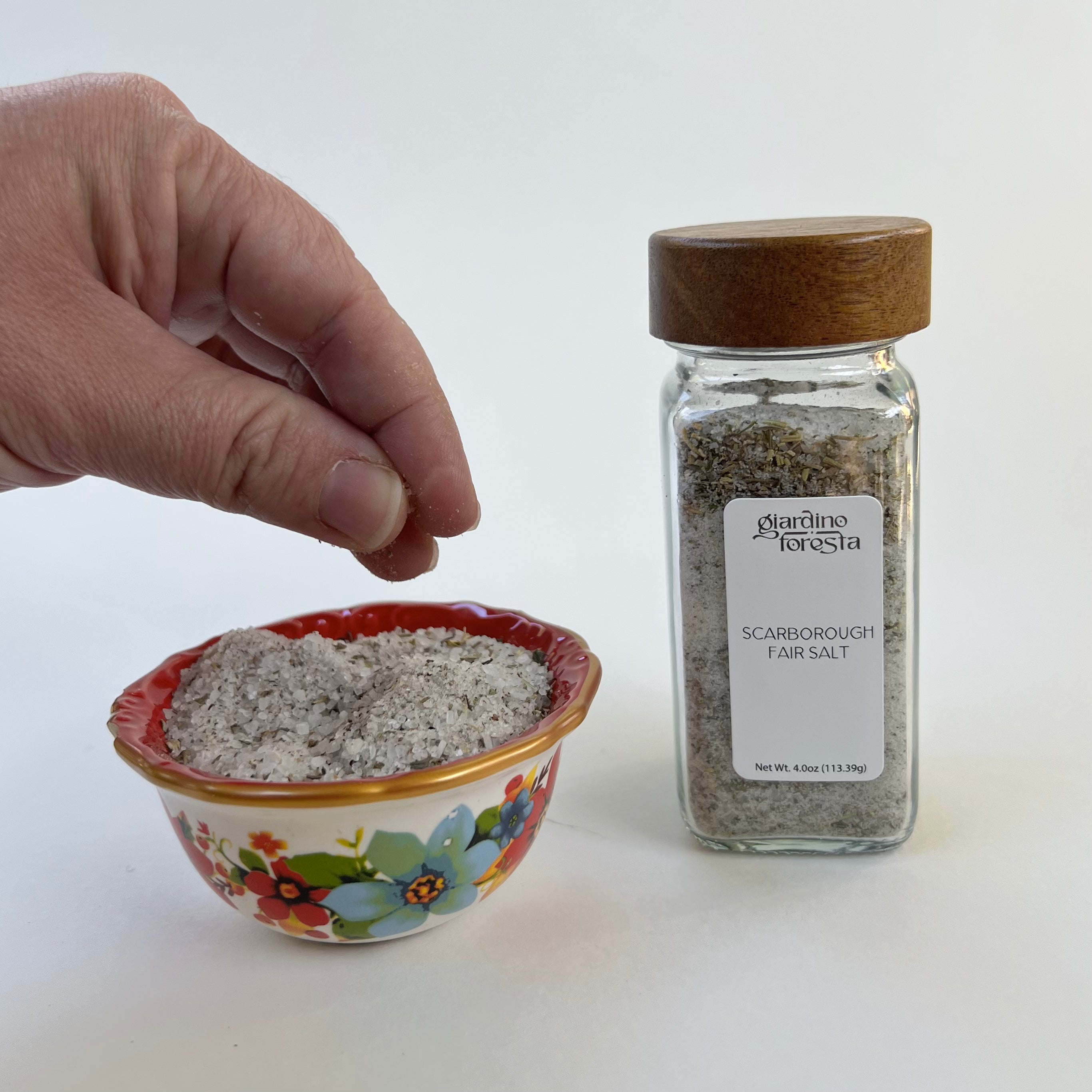 A hand is shown sprinkling salt into a small floral bowl next to a clear glass spice jar labeled Giardino Foresta Scarborough Fair Salt with a wooden lid, on a plain white background.