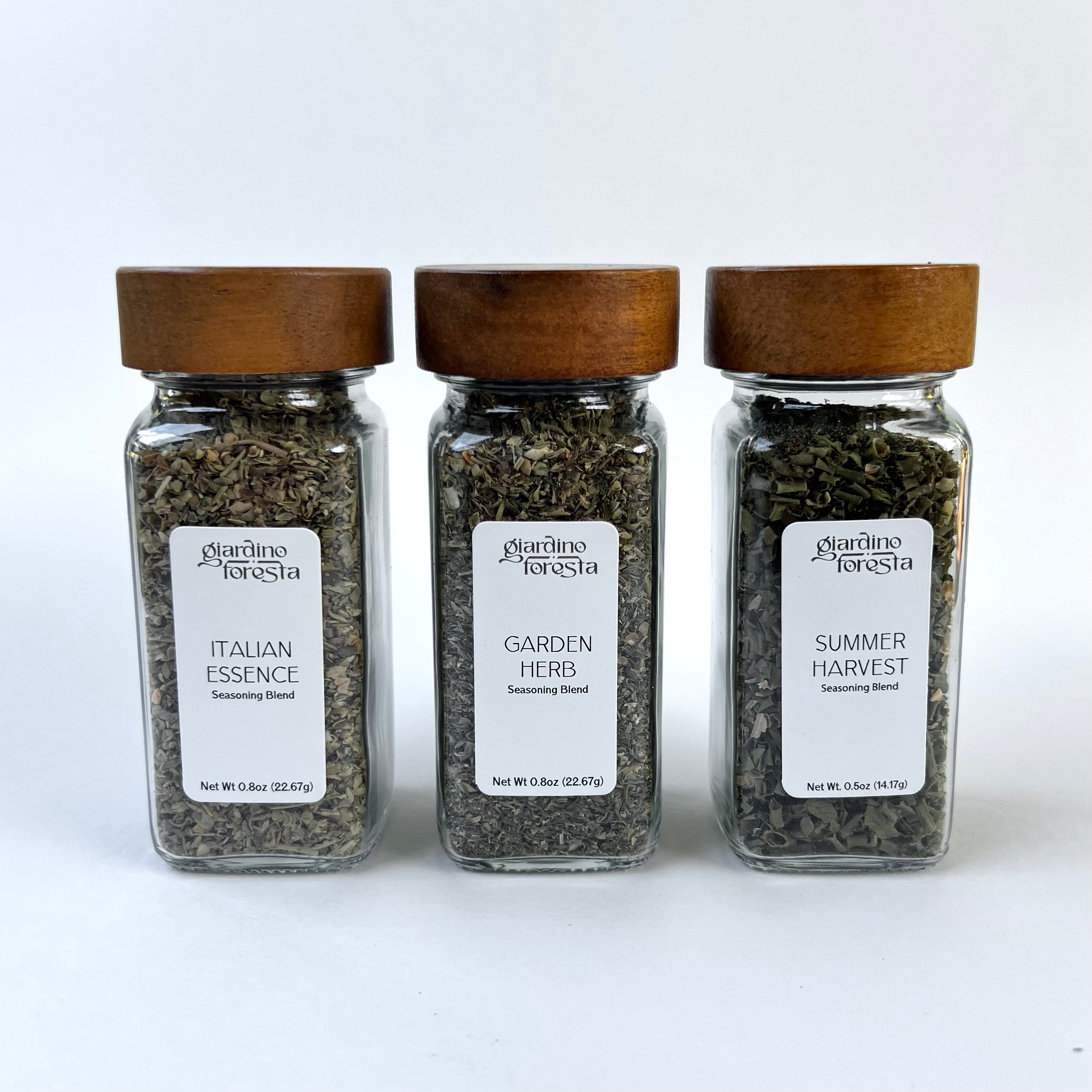 Three clear glass seasoning bottles with wooden lids from Giardino Foresta, arranged side by side against a white background. The labels read Italian Essence, Garden Herb, and Summer Harvest.