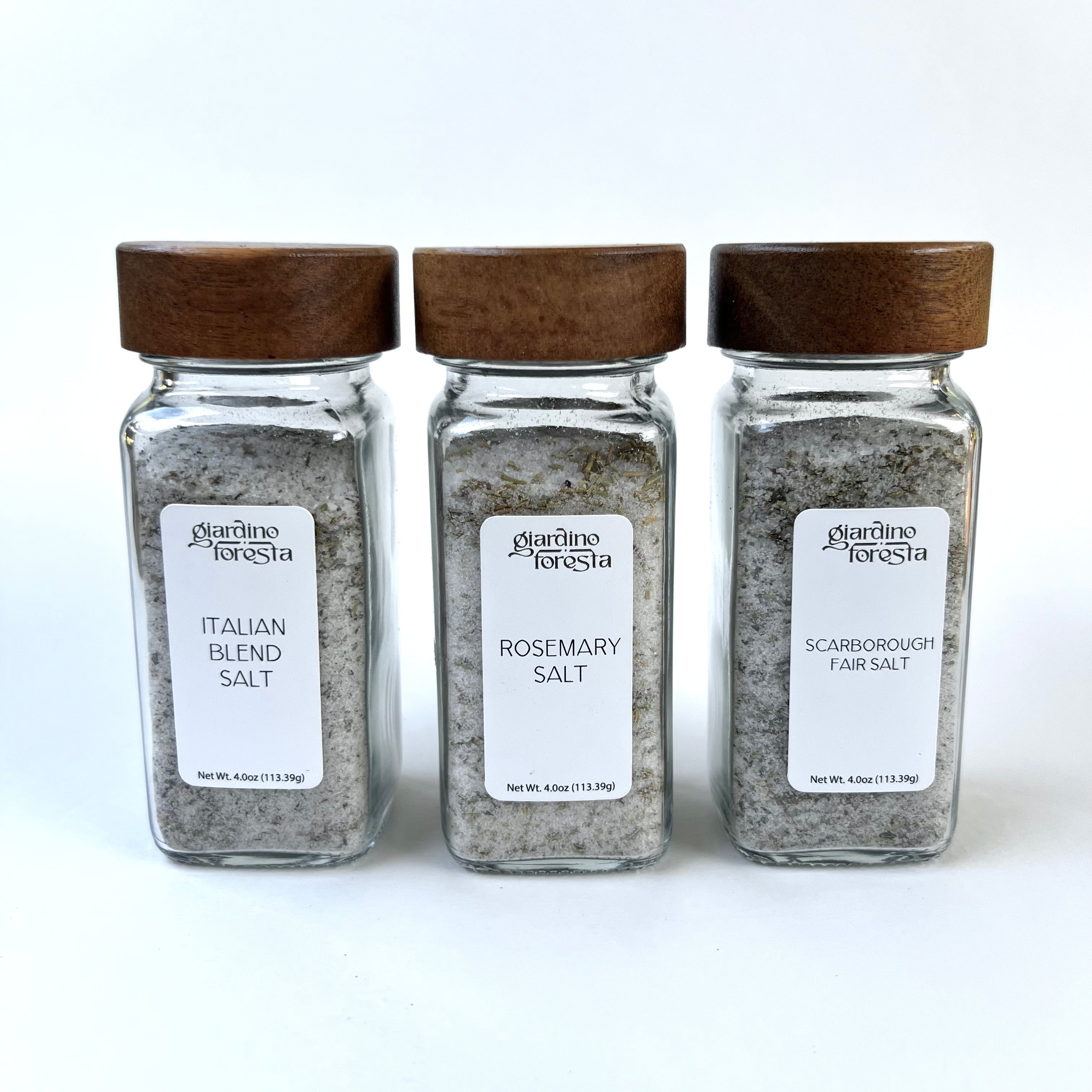 Three glass salt shakers with dark wooden lids from Giardino Foresta, arranged side by side against a white background. The labels read Italian Blend Salt, Rosemary Salt, and Scarborough Fair Salt.