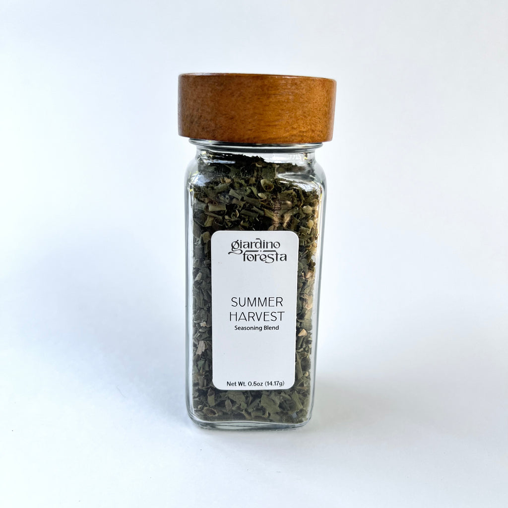 A clear glass spice jar filled with dried green herbs, featuring a white label that reads 'Giardino Foresta Summer Harvest Seasoning Blend' and a medium brown wooden lid, set against a plain white background.