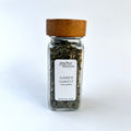 A clear glass spice jar filled with dried green herbs, featuring a white label that reads 'Giardino Foresta Summer Harvest Seasoning Blend' and a medium brown wooden lid, set against a plain white background.