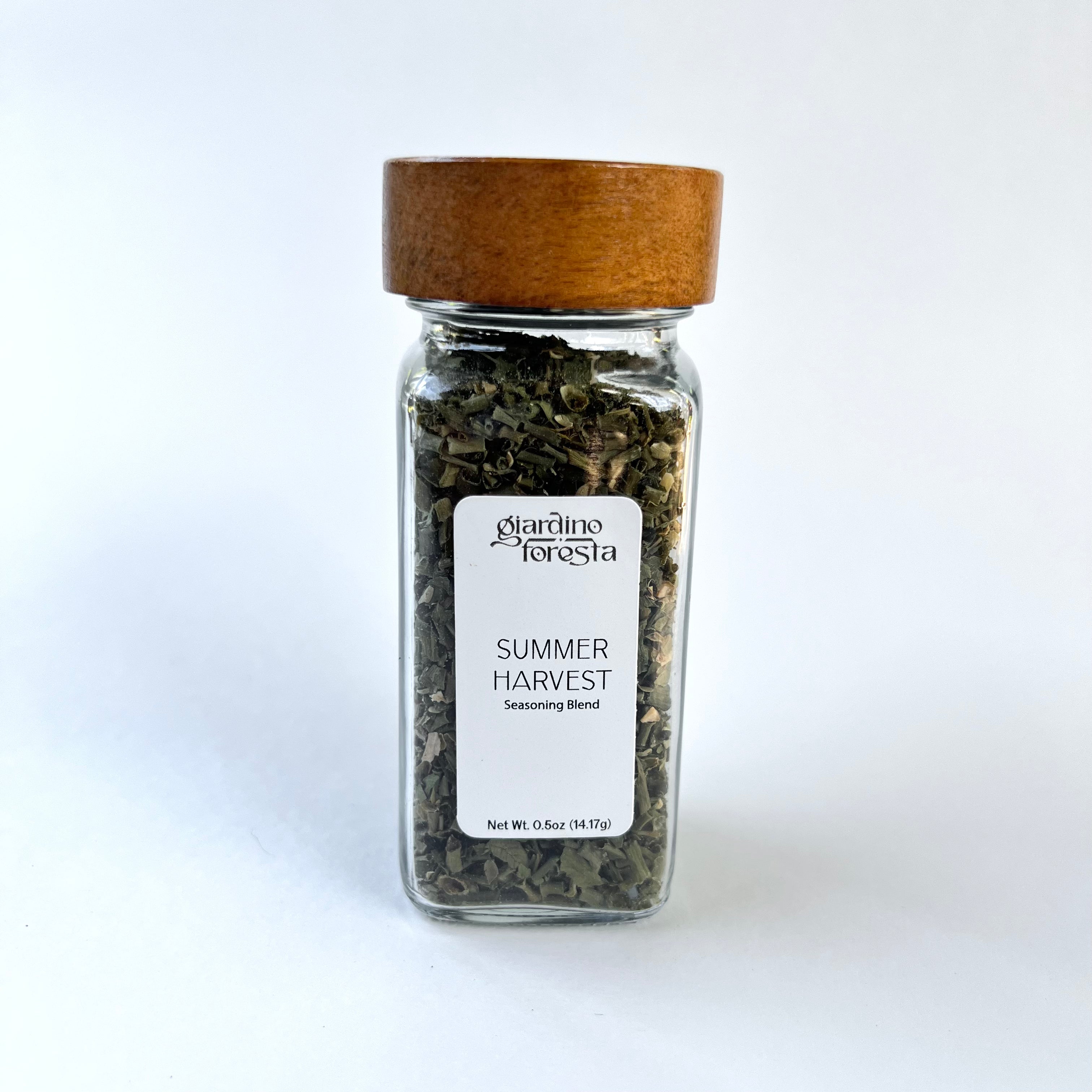 A clear glass spice jar filled with dried green herbs, featuring a white label that reads 'Giardino Foresta Summer Harvest Seasoning Blend' and a medium brown wooden lid, set against a plain white background.