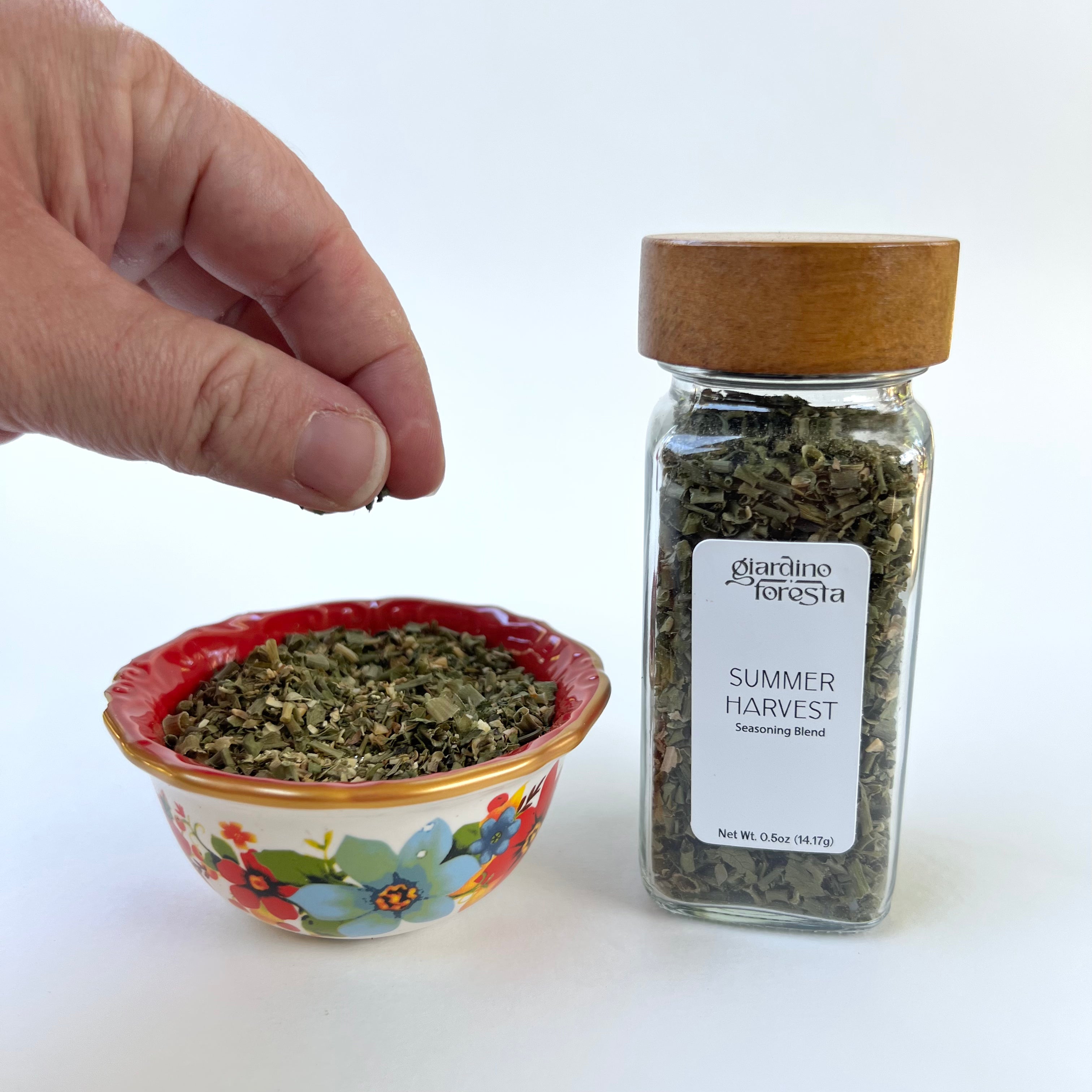 A hand is shown sprinkling dried herbs into a small red and floral bowl next to a glass jar of Giardino Foresta Summer Harvest Seasoning Blend on a white background.