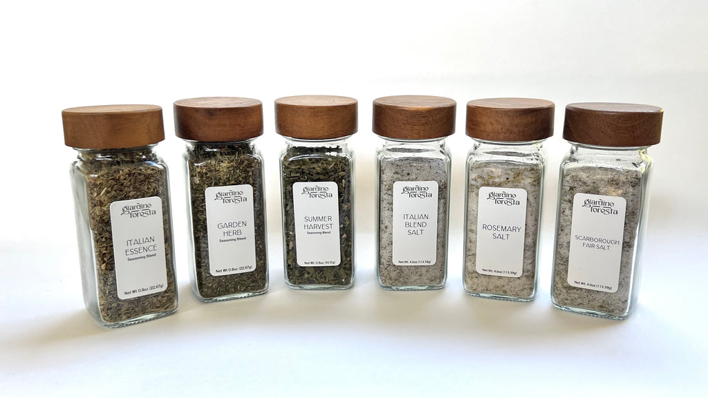 Six clear glass spice jars with wooden lids and white labels from Giardino Foresta, arranged in a row against a white background. The spices include Italian Essence, Garden Herb, Summer Harvest, Italian Herb Salt, Rosemary Salt, and Scarborough Fair Salt.