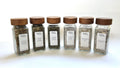 Six clear glass spice jars with wooden lids and white labels from Giardino Foresta, arranged in a row against a white background. The spices include Italian Essence, Garden Herb, Summer Harvest, Italian Herb Salt, Rosemary Salt, and Scarborough Fair Salt.